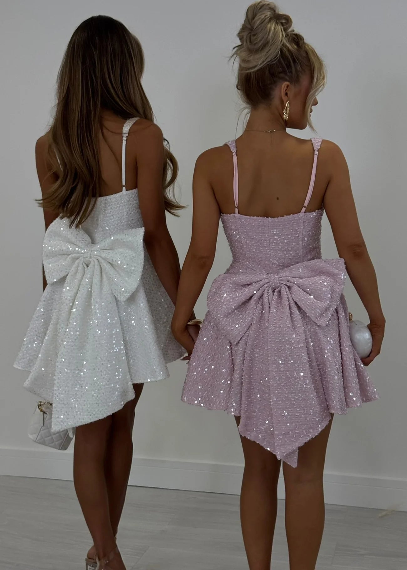 Two women wearing sparkly dresses with large bows, one white and one pink, against a plain background.